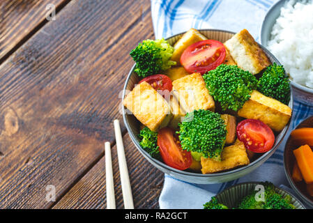 Gegrillter Tofu mit Brokkoli und Tomaten in weiße Schüssel auf Tischdecke mit Stäbchen auf der linken Seite. Stockfoto