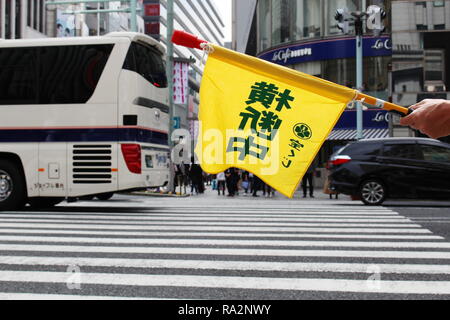 Die Flagge wird von der Grenzwache gehalten, die an einem geschäftigen Übergang in Ginza stationiert ist. 'Crossing' ist auf Japanisch geschrieben. Sanai Dream Center ist im Hintergrund. Stockfoto