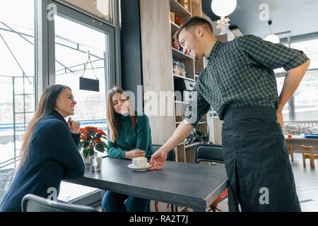 Coffee House, Winter, zwei junge Frauen, die am Tisch sitzen, und eine männliche Barista mit einer Tasse Kaffee zu lächeln und zu sprechen. Stockfoto