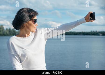 Frau mittleren Alters blogger nimmt Fotos und Videos für Ihr blog auf einem Smartphone. Tag Sommer in der Nähe des Flusses. Stockfoto