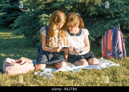 Zwei kleine Mädchen Freunde Schulmädchen lernen sitzen auf einer Wiese im Park. Kinder mit Rucksäcken, Bücher, Hefte. Stockfoto