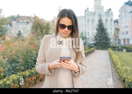 Junge Frau von SMS-Nachrichten auf dem Smartphone zu Fuß in der sonnigen Stadt Straße, Mädchen mit warmen Mantel. Stockfoto