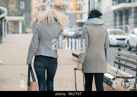 Zwei junge schöne Frauen in warme Kleidung zu Fuß die Straße der Stadt mit einem Reisekoffer, Frauen lachen und reden, Ansicht von der Rückseite. Stockfoto