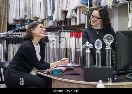 Zwei Frauen arbeiten mit Stoffe für Vorhänge, Polstermöbel, lächelnde Frauen wählen Sie Strukturen mit Computer. Workplace Designer, Verkäufer von Dekostoffe Stockfoto
