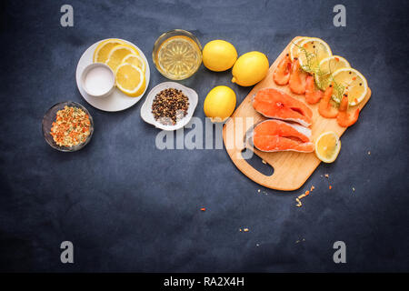 Roher Fisch mit Gewürzen. Auf einem grauen Tischplatte läuft ein hölzernes Brett auf dem Lachs rohe Garnelen Zitronenscheiben Pfeffer Öl würzen. Stockfoto