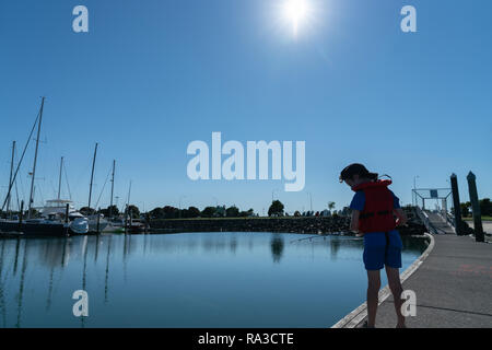 Kleiner Junge schaut in roter Schwimmweste Angeln vom Steg der Marina an sonnigen ruhigen Morgen Sonne Silhouette Stockfoto