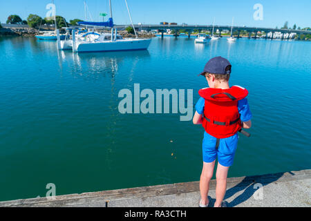 Kleiner Junge von hinten in rot Rettungsweste Angeln vom Steg der Marina an sonnigen ruhigen Morgen mit angelegten Boote und Tauranga Bridge im Abstand Hafen Stockfoto