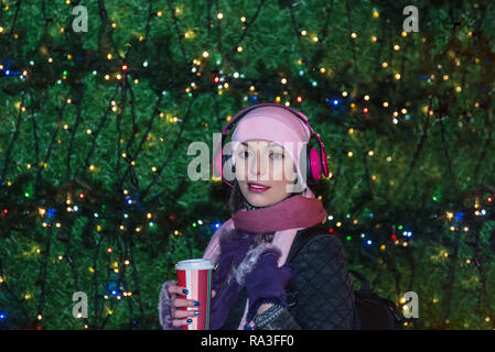 Schöne Frau in Beanie-mütze mit Kaffee zum Mitnehmen und Kopfhörer stellt sich vor Weihnachten Dekoration leuchten. Stockfoto