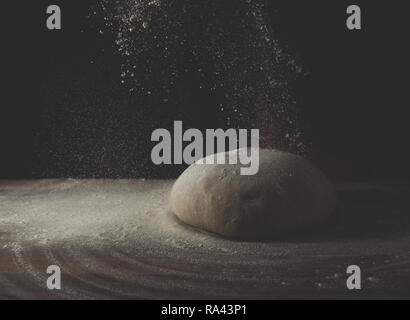 Den Teig in die Form einer Schüssel liegt auf einer hölzernen backen Tabelle auf einem dunklen schwarzen Hintergrund. Mit Mehl bestreut. Seitenansicht, horizontale Foto Stockfoto