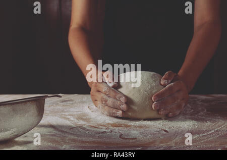Der Teig durch weibliche Hände auf einer hölzernen backen Tabelle. Dunkler schwarzer Hintergrund mit Mehl. Seitenansicht, horizontale Foto Stockfoto