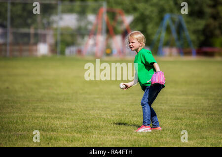 Ein sechs Jahre altes Mädchen in Jeans und T-Shirt und rosa Handschuh fertig, einen Baseball auf grüner Sommer Gras zu werfen Stockfoto