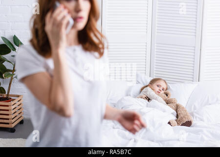 Selektiver Fokus der Mutter ruft Arzt per Smartphone, kranken Tochter im Bett Stockfoto