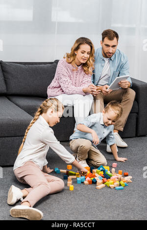 Die Eltern sitzen auf einem Sofa mit digitalen Tablet und spielende Kinder mit bunten Holzklötzen auf dem Fußboden in der Wohnung Stockfoto