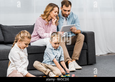Die Eltern sitzen auf dem Sofa und Verwendung digitaler tablet, während die Kinder spielen mit bunten Holzklötzen auf Fußboden im Wohnzimmer Stockfoto