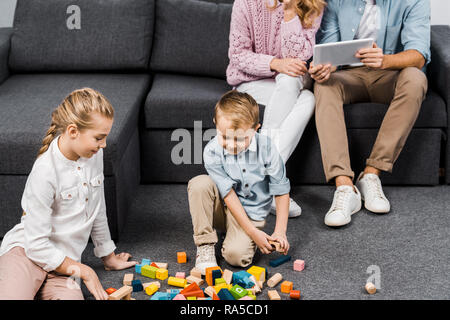 7/8-Ansicht der Eltern sitzen auf dem Sofa und Verwendung digitaler tablet, während die Kinder spielen mit bunten Holzklötzen auf Fußboden im Wohnzimmer Stockfoto