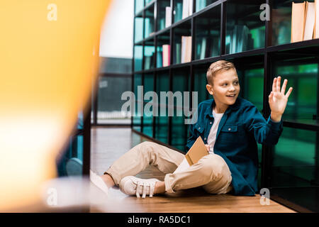 Nett lächelnden Jungen holding Buch und winkende Hand beim Sitzen auf dem Boden in der Bibliothek Stockfoto