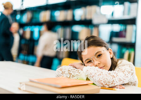 Gelangweilte Schulmädchen in Brillen lehnte sich an den Tisch mit Büchern und Kamera in Bibliothek Stockfoto