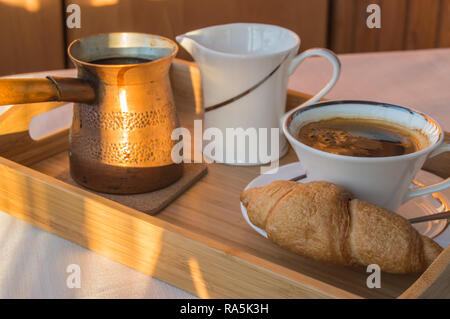 Das Frühstück wird auf einer Holz- Fach mit Kupfer Töpfe serviert, eine Tasse Kaffee und Croissants. Sonnenlicht auf dem offenen Balkon am Morgen. Stockfoto