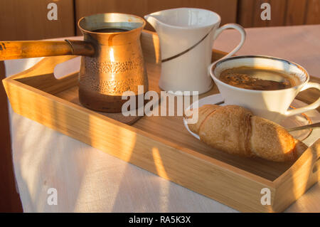 Das Frühstück wird auf einer Holz- Fach mit Kupfer Töpfe serviert, eine Tasse Kaffee und Croissants. Sonnenlicht auf dem offenen Balkon am Morgen. Stockfoto