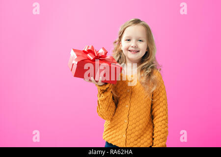 Glückliches Kind Mädchen in einem warmen gestrickten Pullover Holding eine Box mit einem Geschenk, auf rosa Hintergrund. Das Konzept von Geben und Empfangen von Geschenken. Stockfoto