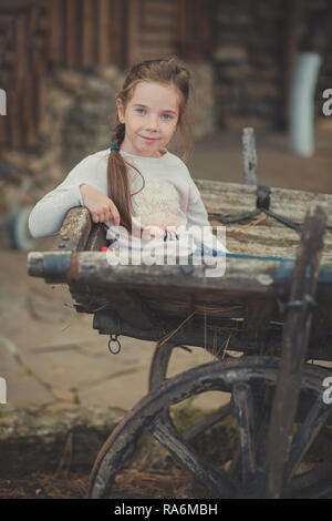 Baby junge Mädchen mit blauen Augen mit brunnette Zopf Haar tragen weiße Kleid Shirt und Posieren auf hölzernen old style retro Wagen Warenkorb trundle Posing suchen Stockfoto