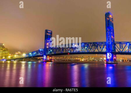 Die Jacksonville Florida Hauptbrücke blau lackiert und bei Nacht beleuchtet mit einer Überlegung über den St. Johns Fluss und die Skyline im Hintergrund Stockfoto