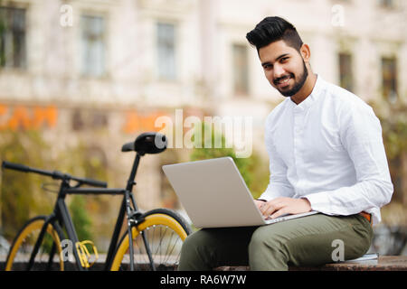 Hübscher junger Geschäftsmann im Anzug Arbeiten am Laptop gekleidet beim Sitzen im Freien Stockfoto