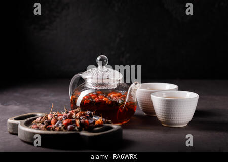 Eine Teekanne aus Glas mit einem frisch gebrühten Vitamin rose Drink und zwei Schüsseln. Hagebutte Tee. Vitamin Drink auf einem dunklen Hintergrund. Close-up. Stockfoto