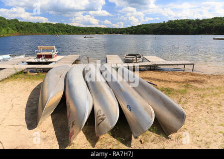 Eine Reihe von silbernen Kanus sind am Strand eines Sees im Wald während des Sommercamps aufgereiht. Stockfoto