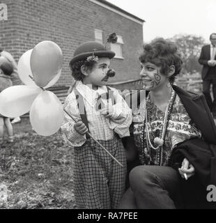 1965, Dorffest, die Mutter mit dem Wort "Liebe" auf Ihrem Gesicht gemalt, mit ihr, ein kleiner Junge in Fancy Dress, trug einen Hut und eine Nase wie ein Clown und Halten der Ballons, England, UK. Stockfoto