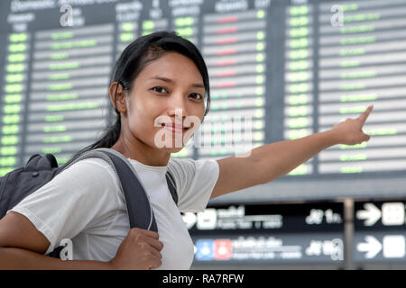 Reisen Frau mit einem Rucksack auf dem Rücken, die ein Schild mit den Flugverbindungen. Die asiatische Mädchen mit dem Gepäck ist unter Vorstand am Flughafen. Stockfoto