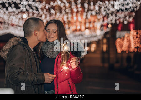 Junge glückliches Paar Spaß mit Wunderkerzen im Winter city night. Der junge Mann Küsse und Umarmungen seiner Freundin Stockfoto