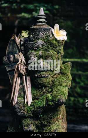 Stein Skulptur eines Gottes in einem Garten auf Bali Indonesien mit Frangipani Blume große Augen eine Krone und Zähne Stockfoto