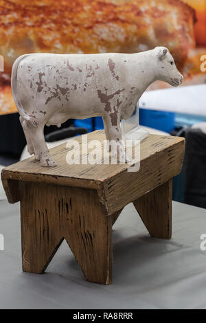 Rustic weiß lackiert Kuh statue sitzend auf einem Verwitterten braunes Holz Bank auf einem Tisch Stockfoto