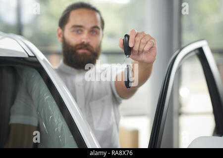 Nahaufnahme der Autoschlüssel. Windschutzscheibe von weißes Auto mit geöffneter Tür. Schöne bärtige hinter Mann stand. Client Autohaus im weißen Hemd Holding auto Schlüssel in der Hand. Stockfoto