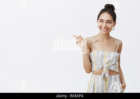 Studio shot von Happy elegante Hündin mit gebräunter Haut und Mole unter der Lippe, freudig lächelnd während zeigen und den Blick nach links, genießen die große Zeit auf formale Abend zu ihr b-Tag Stockfoto