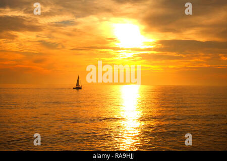 Ein Segelboot mit drei Menschen in der Ferne in einer ruhigen orange leuchtende nahtlose Meer Silhouette die Sonne durch die Wolken und Cas scheint Stockfoto