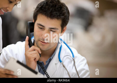 Junge männliche Arzt am Telefon, während in der Zwischenablage, die eine junge Krankenschwester ihm zeigt. Stockfoto