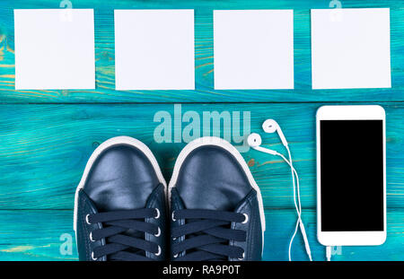 Ansicht von oben von Sport Schuhe per Handy mit isolierten Bildschirm und In-Ear-Kopfhörer und weiße Leere leere Blätter auf schwarz Holztisch. Emp Stockfoto