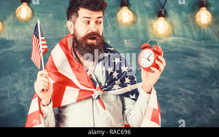 Mann mit Bart auf überrascht Gesicht hält Flagge der USA und Uhr, schwarzes Brett für den Hintergrund. Amerikanische Lehrer mit amerikanischen Fahnen hält Wecker. Amerikanischen Bildungssystem Konzept. Stockfoto