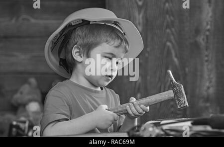 Kid boy nail Hämmern in Holzbrett. Kind in Helm cute Spielen als Bauherr oder Reparatur, Instandsetzung oder Basteln. Kleinkind bei Besetzt Gesicht spielt zu Hause in der Werkstatt. Basteln Konzept. Stockfoto