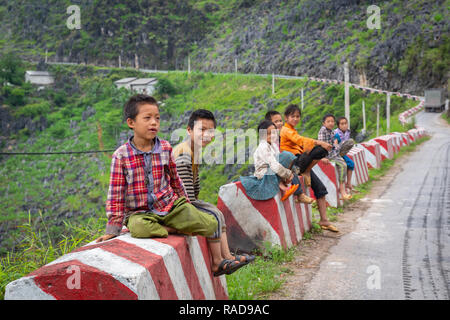 Gruppe junger vietnamesischer Kinder sitzen auf einem gestreiften Straßenrand konkrete Schutzgeländer in Ha Giang Loop, Provinz Ha Giang, Vietnam, Asien Stockfoto