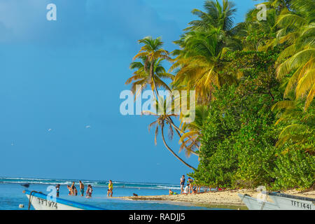 Pigeon Point, Tobago, Karibik. Familie oder eine Gruppe von Menschen, am Strand entlang schlendern mit wiegenden Palmen, strahlend blauen Himmel und das blaue Meer. Landschaft Stockfoto