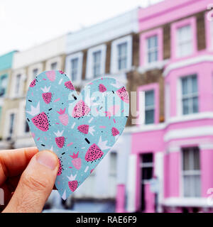 Nahaufnahme von einem kaukasischen Mann hält ein Herz Ausschnitt auf ein buntes Papier, vor einer Reihe von bunten Häusern in der beliebten Portobello Road in London, Stockfoto