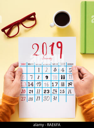 Hohe Betrachtungswinkel von einem kaukasischen Mann beobachten ein Kalender von 2019 an einem gelben Tisch sitzen neben eine Tasse Kaffee, ein Paar rote Brille und einem Gree Stockfoto