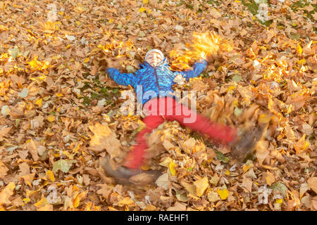 Knabenkind, das in heruntergefallenen Blättern spielt, indem er seine Arme und Beine auf dem Boden liegend bewegt. Stockfoto