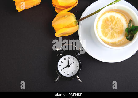 Schwarz Wecker in der Nähe von Bouquet gelbe Tulpen und Schale der Zitrone Tee auf schwarzen Hintergrund. Flach. Mutter oder Frau Tag. Grußkarte. Guten Morgen bre Stockfoto