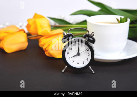 Schwarz Wecker in der Nähe von Bouquet gelbe Tulpen und Schale der Zitrone Tee auf schwarzen Hintergrund. Mutter oder Frau Tag. Grußkarte. Guten Morgen Frühstück. Co Stockfoto