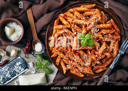 Lecker Penne Arrabiata auf einem Steingut Teller mit frischem Basilikum und geriebenem Pecorino auf einem rustikalen Tisch mit braunen Tuch, Gabel und Zutaten Stockfoto