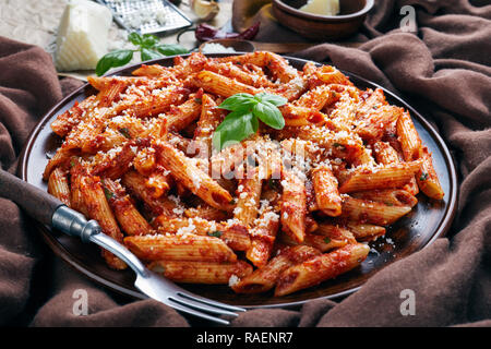 Nahaufnahme der klassische italienische Penne Arrabiata mit Hot Tomato Sauce auf einem Ton Platte mit frischem Basilikum und geriebener Ziegenkäse auf einem rustikalen Tisch mit Braun Stockfoto